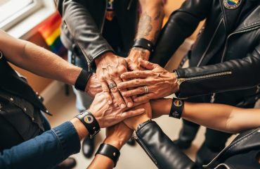 Overhead close-up of diverse LGBTQ leather community members stacking hands, subtle leather cuffs and rings visible, with a softly blurred community room and pride colors in the background.